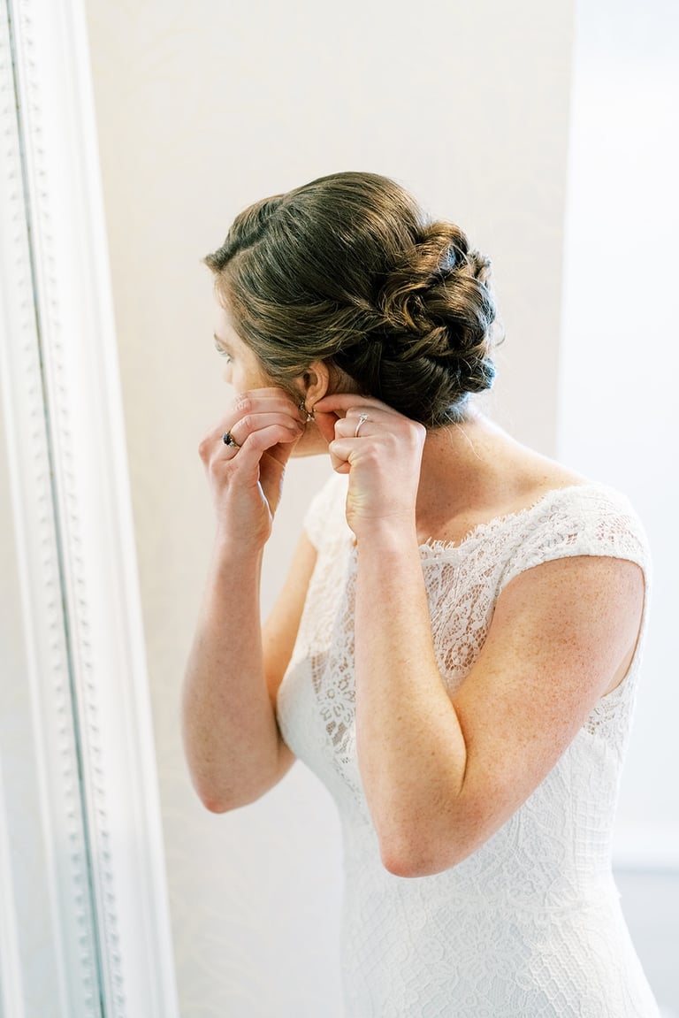 A brunette bride with a freshly completed low updo hairstyle putting on her earrings.