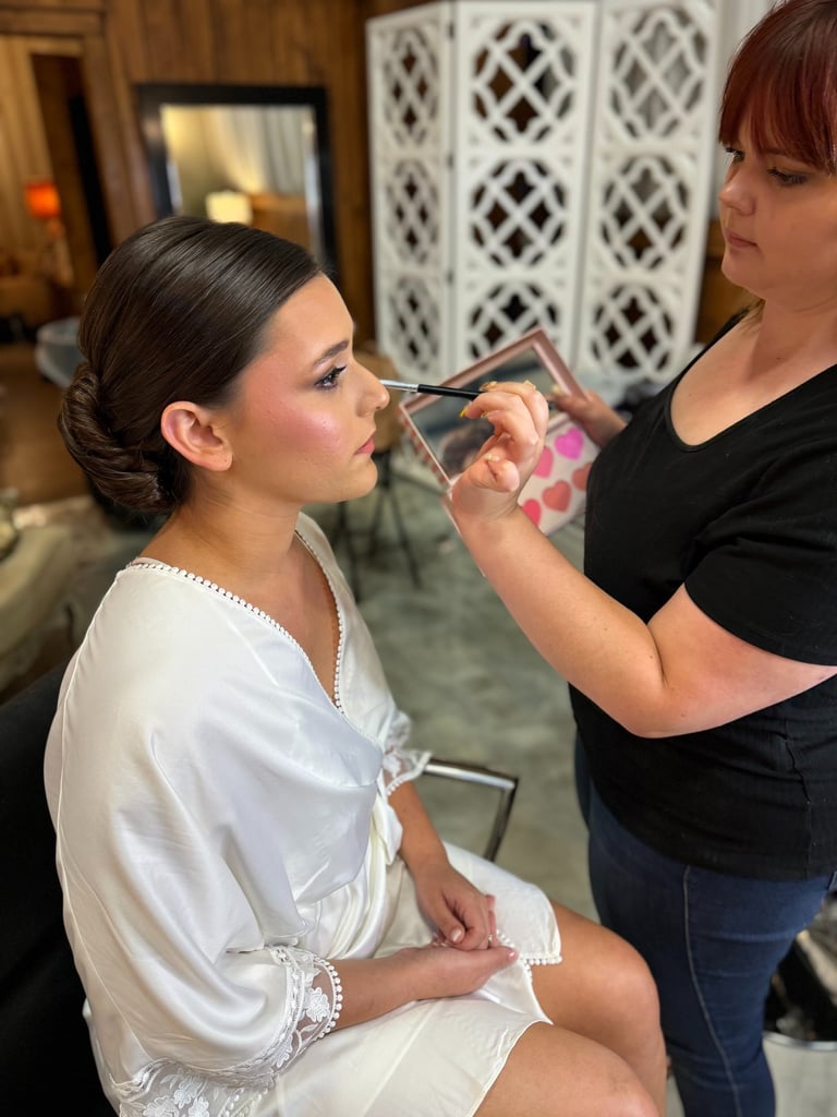 Bride in a satin robe with sleek low bun as a makeup artist perfects eyeliner, highlighting wedding hair and makeup.
