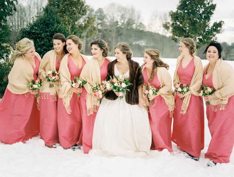 Bride in fur wrap with bridesmaids in coral dresses smiling in the snow, showing elegant wedding hair and natural makeup.