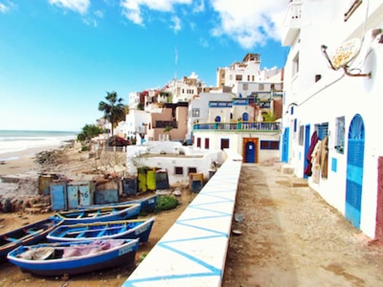 Blue fishing boats docked on a sandy beach next to white coastal buildings in Taghazout, Morocco.