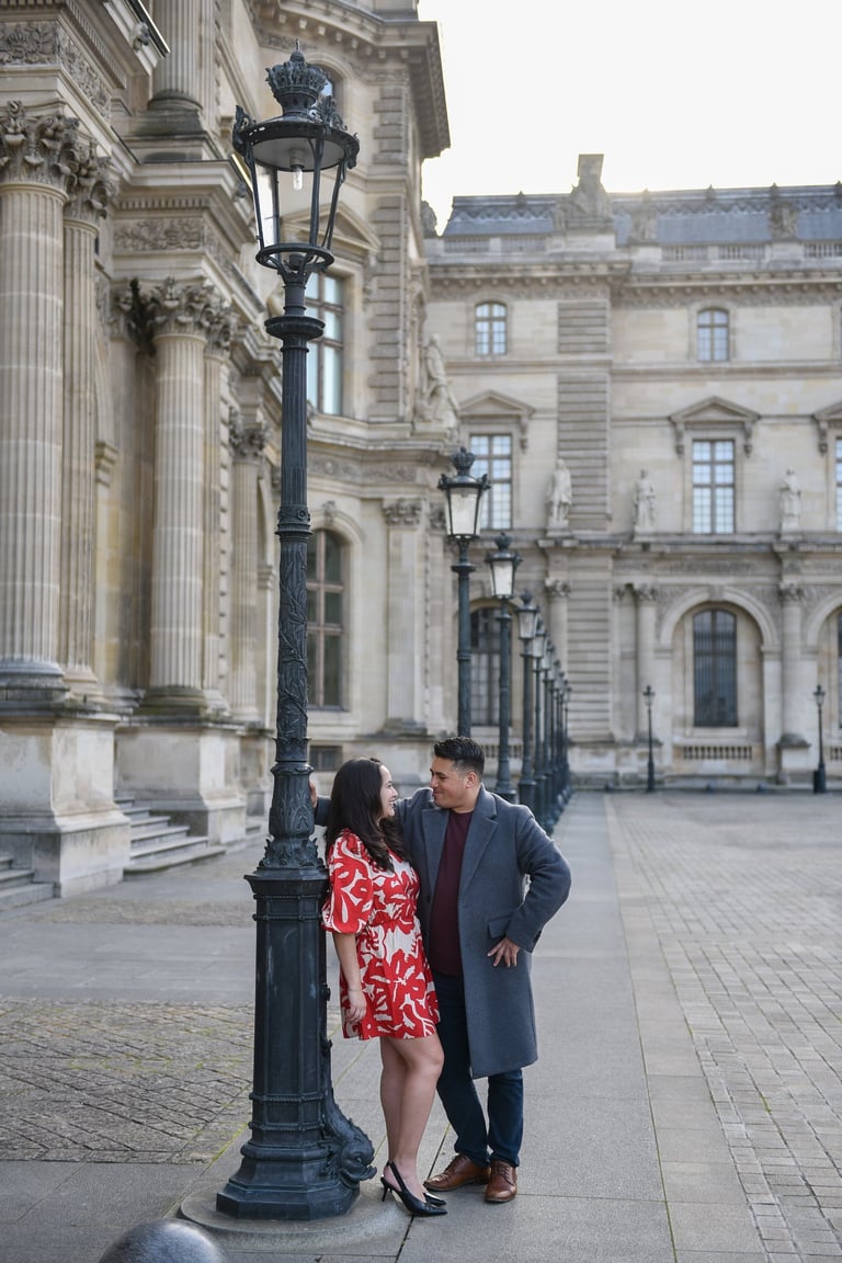 Un couple d'amoureux posent contre un lampadaire dans la cour du Louvre à Paris. On voit le musée en arrière plan