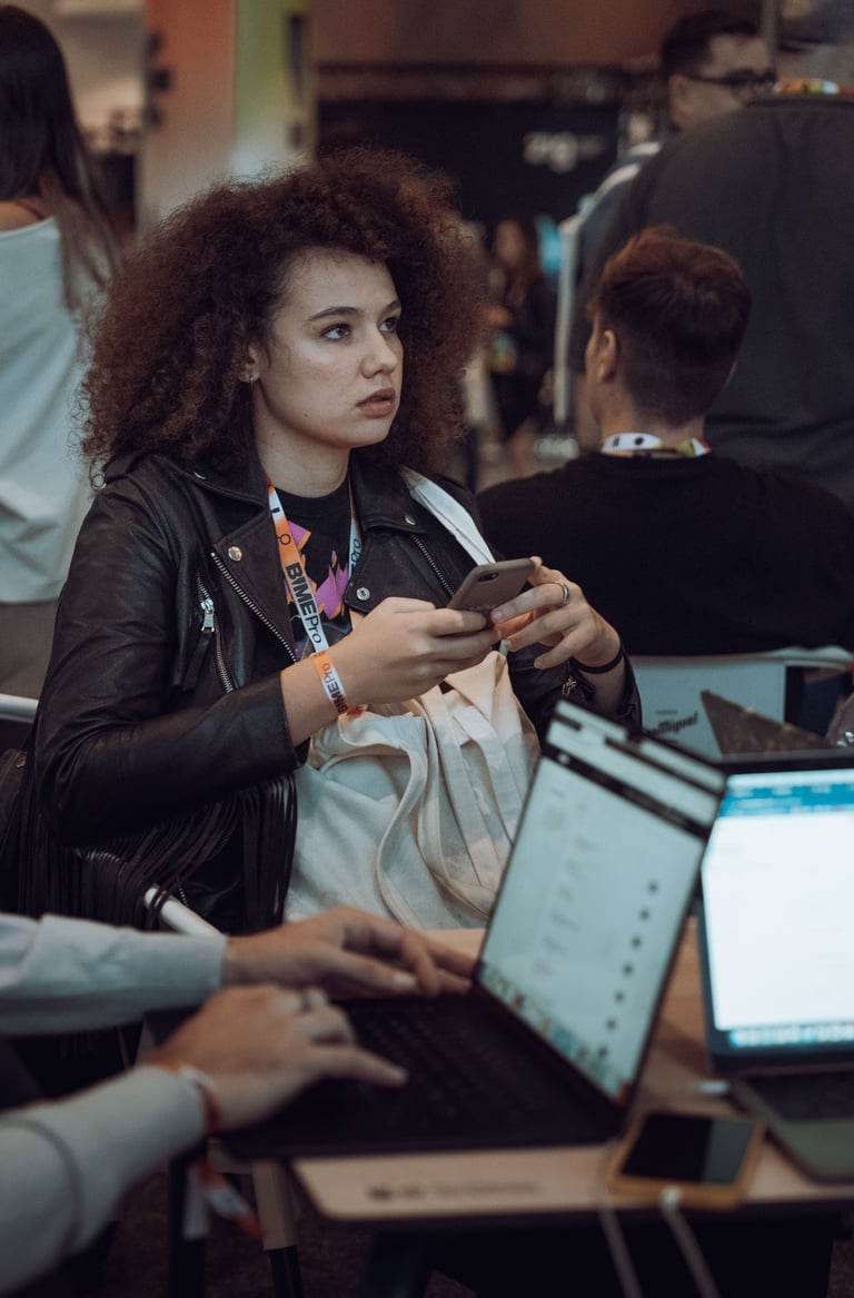 Woman with curly hair wearing a BIME Pro lanyard, looking at her phone at BIME Bilbao. Laptops and other attendees nearby. Ne