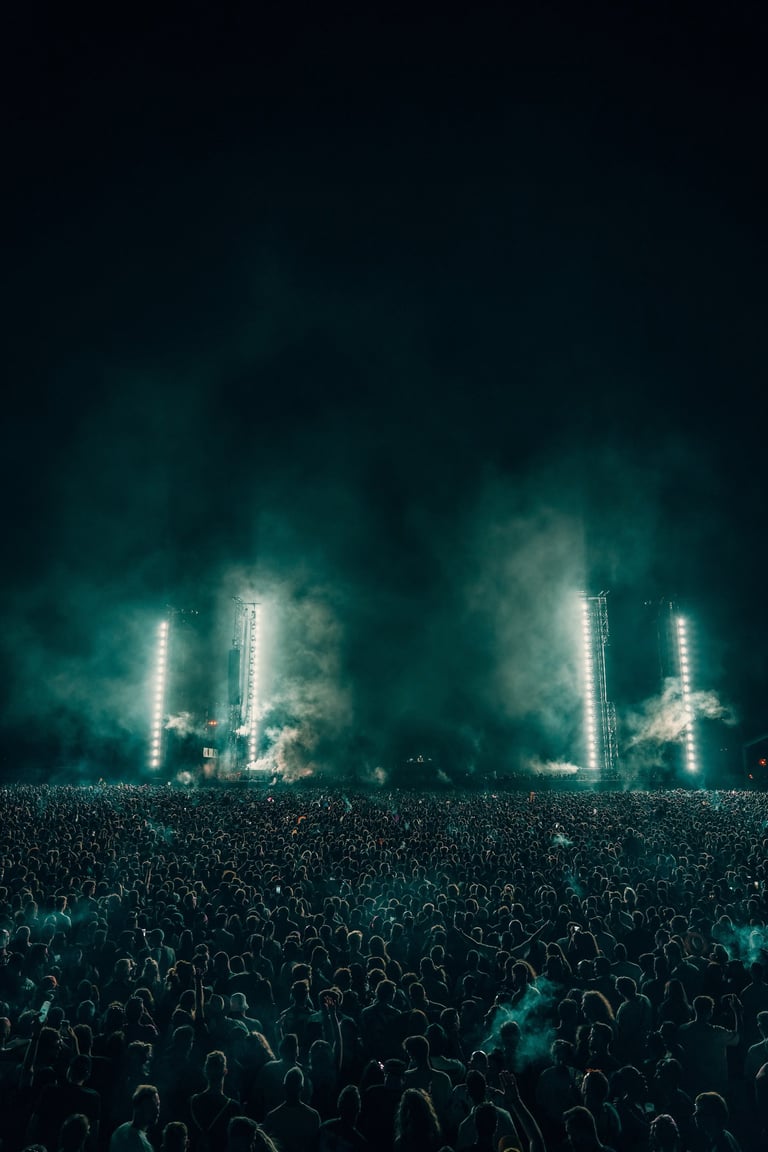 Large crowd at a night concert with bright stage lights and smoke effects. Dark sky above. Energetic music festival scene