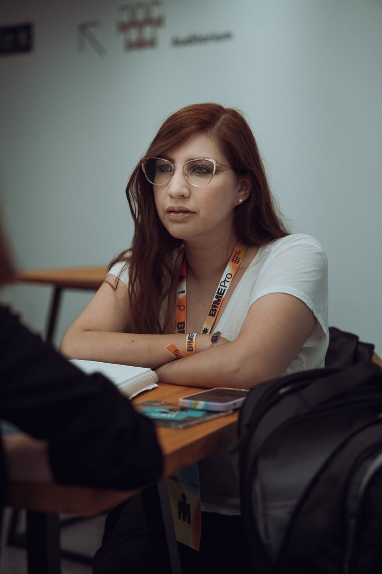 Woman with red hair and glasses wearing a BIME Pro lanyard, sitting at a table with a notebook and phone at BIME Bilbao. Atte