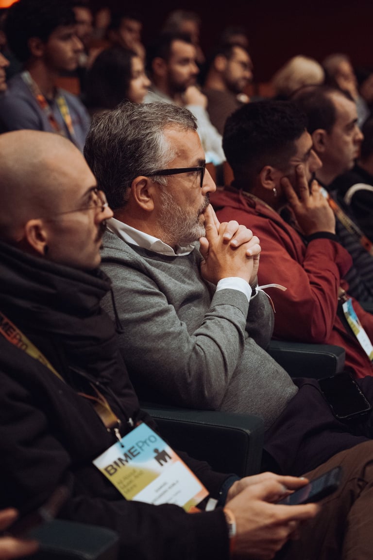 Attendees watching a BIME Bilbao event. Focus on a man with glasses and a grey sweater, hands clasped. Indoor conference sett