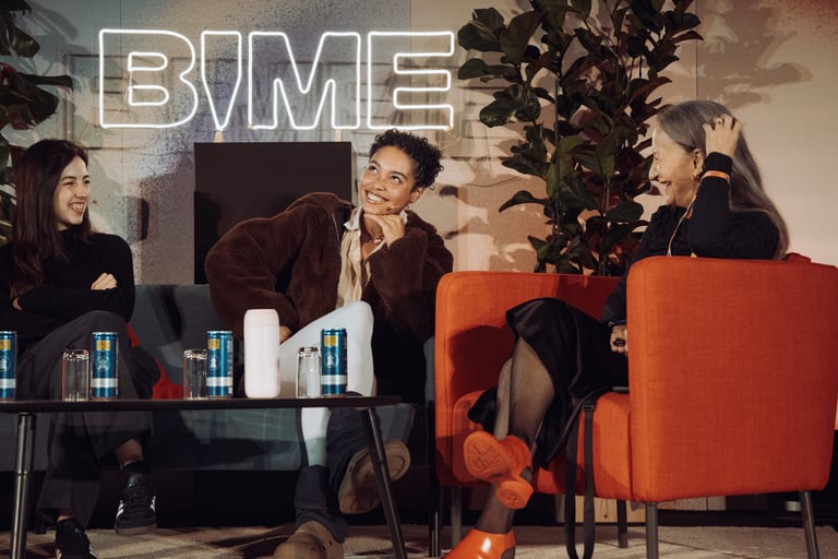 Three women in a relaxed conversation at BIME Bilbao. Neon "BIME" sign and plants in the background. Informal discussion sett