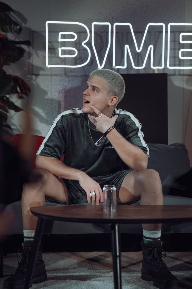 Young man with bleached hair sitting at a table at BIME Bilbao, looking upwards. Neon "BIME" sign in the background. Casual m