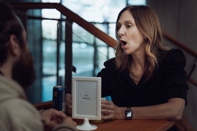 Woman with long brown hair talking animatedly at a table with a BIME Bilbao name card and a drink. Another person partially v