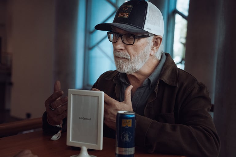 Older man with glasses and a cap gesturing while seated at a table with a BIME Bilbao name card and a drink. Indoor meeting