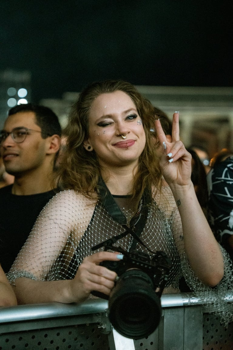 Woman winking and making a peace sign while holding a camera at an event. Person with glasses in the background. Dark setting