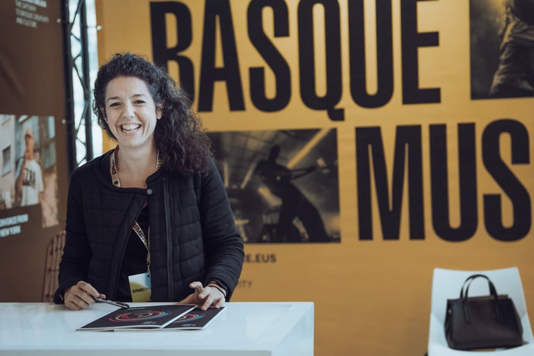 Smiling woman with a BIME Bilbao lanyard standing behind a desk with "BASQUE MUSIC" backdrop. Promoting Basque music at the e