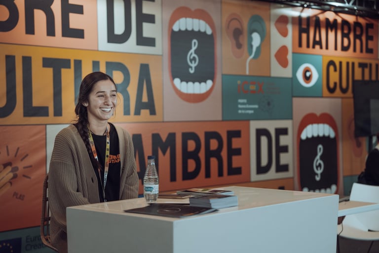 Smiling woman with a BIME Bilbao lanyard sitting at a white desk with promotional materials. Colorful "HAMBRE DE CULTURA" bac