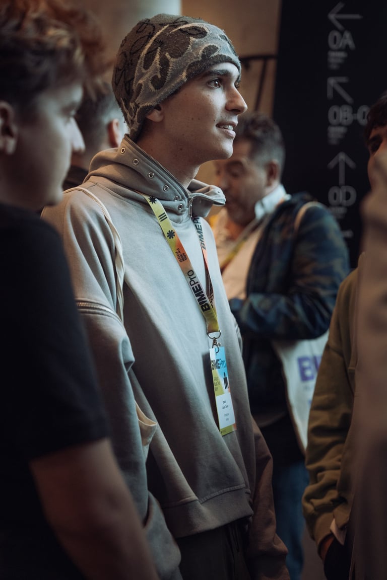 Young man wearing a BIME Pro lanyard at BIME Bilbao, smiling and looking to the side. Surrounded by other attendees. Conferen