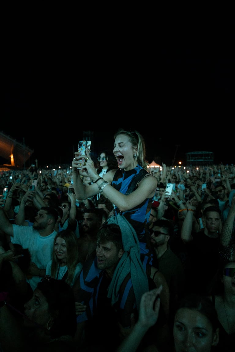 Excited woman on someone's shoulders, filming a concert. Energetic crowd with phones raised in the dark. Live music atmospher