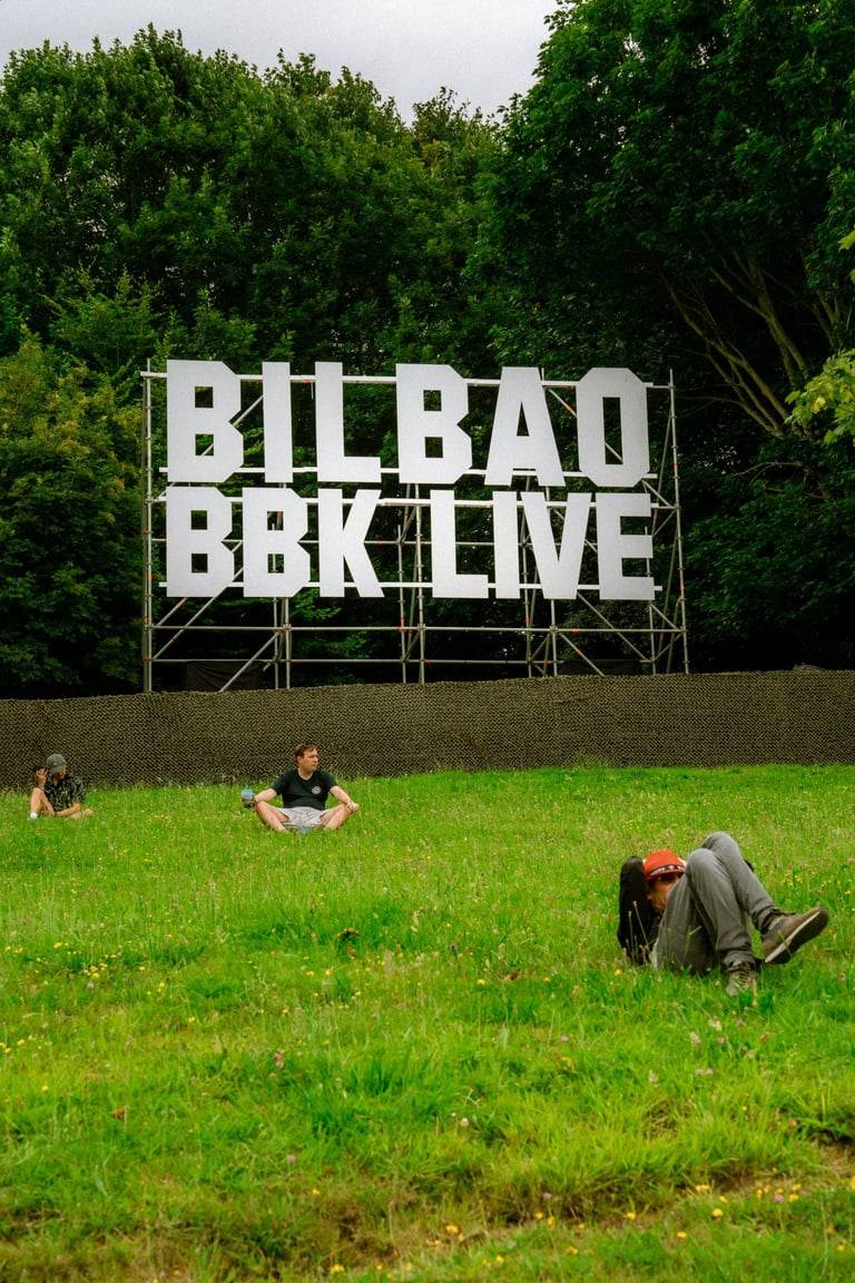 People relaxing on the grass in front of a large "BILBAO BBK LIVE" sign. Green trees in the background. Festival entrance