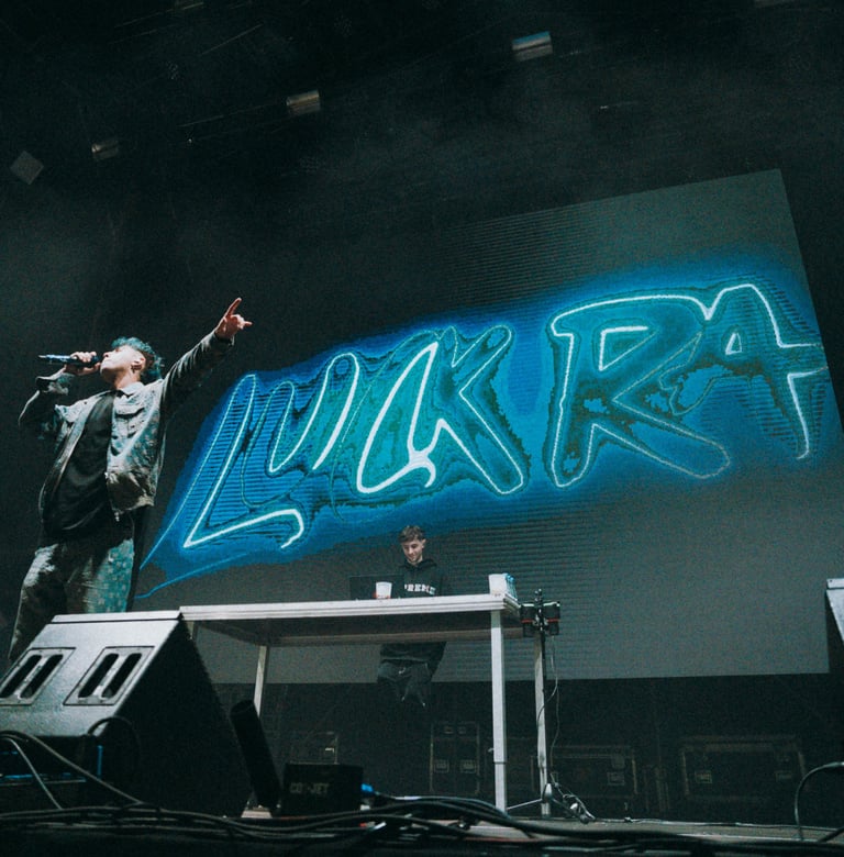 Luck Ra performing live at Boombastic Festival in Bilbao. Stage with neon "Luck Ra" backdrop. Energetic concert atmosphere