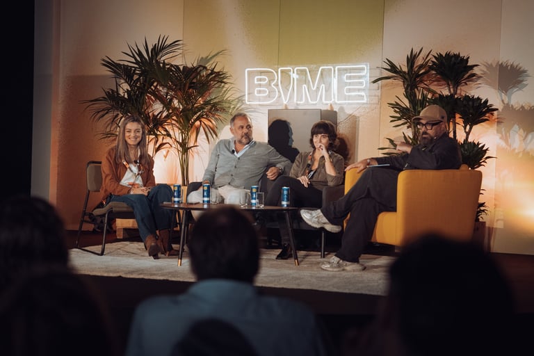 Panel discussion at BIME Bilbao. Four speakers seated on a stage with a neon "BIME" sign and palm trees. Audience in the fore
