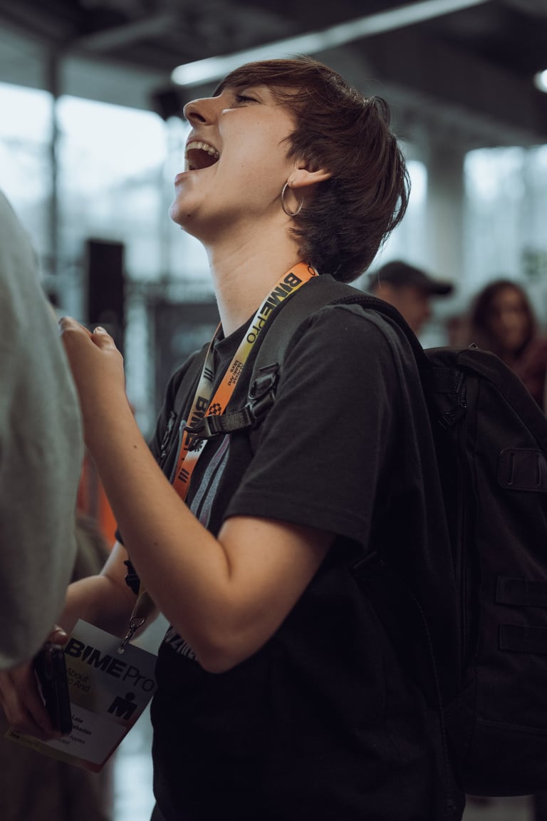 Young person with short brown hair, wearing a BIME Bilbao lanyard and a backpack, laughing with their mouth open. Indoor sett