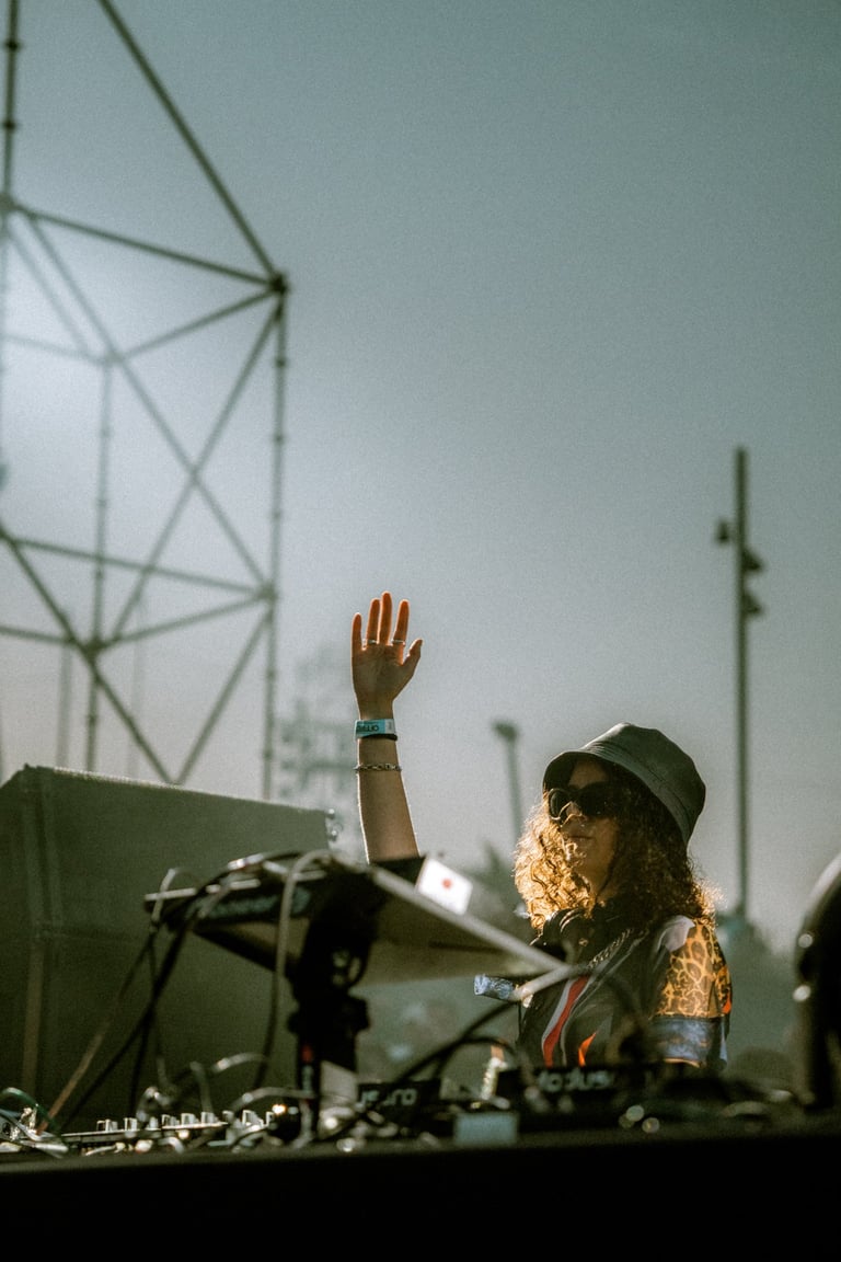 Female DJ with curly hair and sunglasses performing at an outdoor music event. Hand raised in the air. Stage and sky backgrou