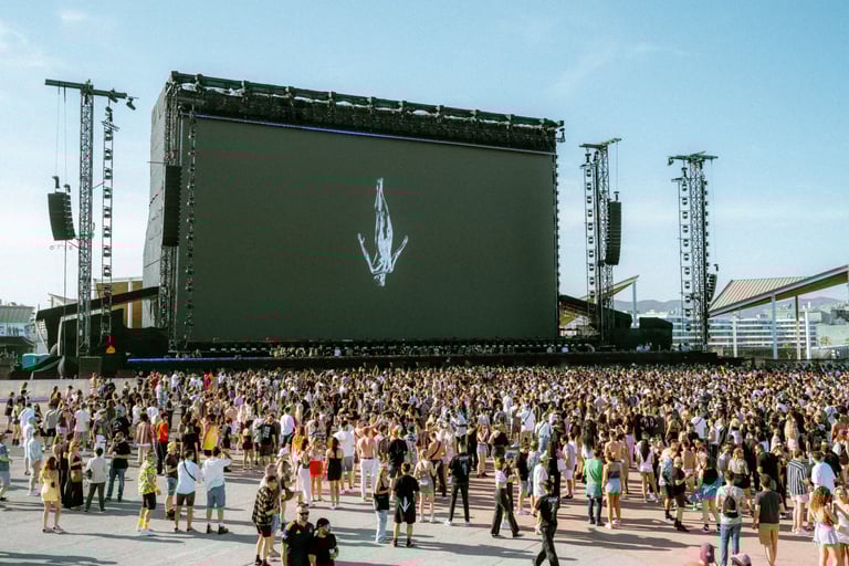 Large crowd gathered at a daytime music festival, facing a massive stage with a screen displaying an image. Afterlife