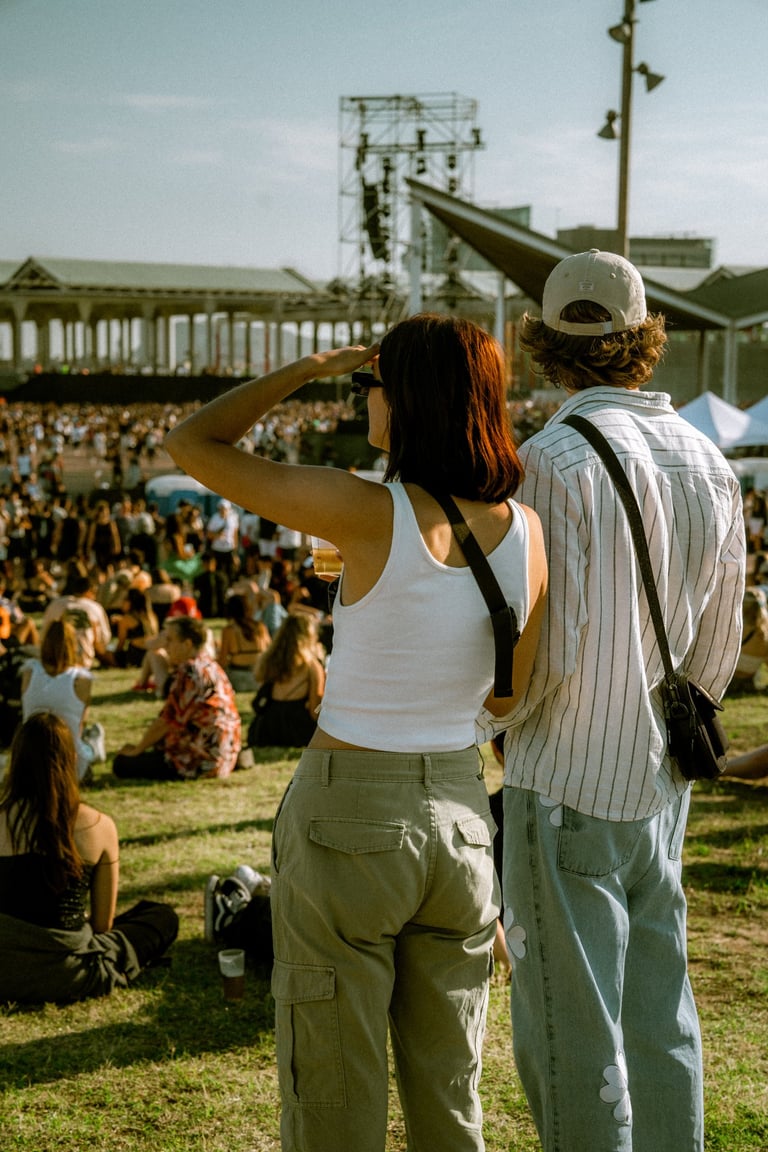 Couple watching a festival stage from a grassy field. Large crowd of people enjoying an outdoor event under a sunny sky.