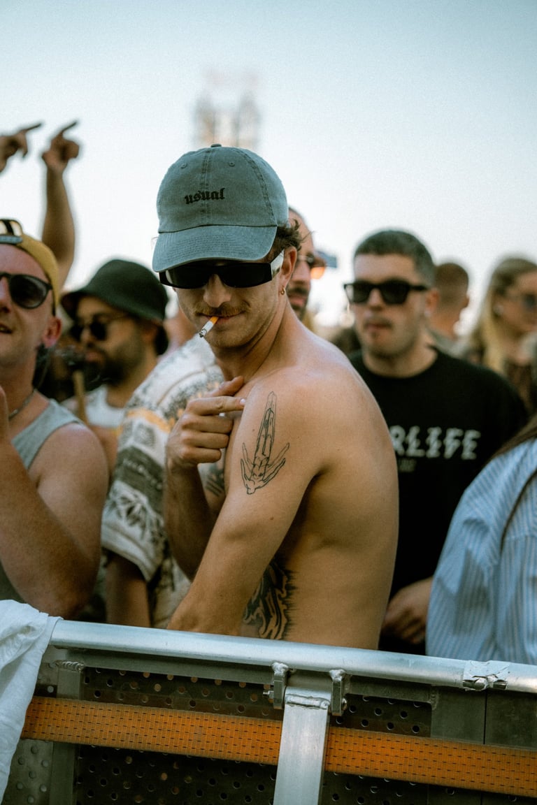 Shirtless man with tattoos, sunglasses, and a cap smoking in a crowd at an outdoor event. People around him. Casual atmospher