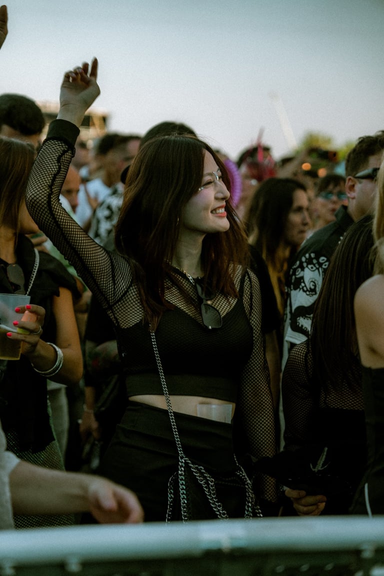 Smiling woman with glitter on her face dancing in a crowd at an outdoor festival. Arm raised in the air. Festive atmosphere