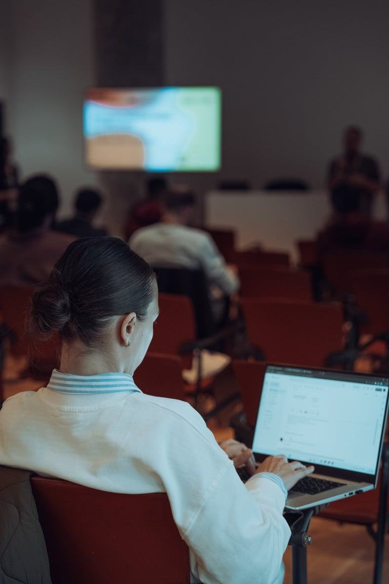 Attendee with a laptop at BIME Bilbao, watching a presentation on a screen. Red chairs in a conference room. Learning and eng