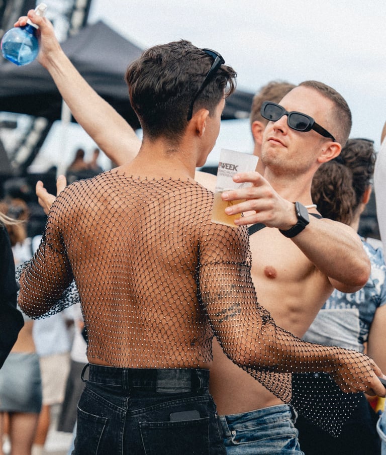 Two men at an outdoor event, one shirtless with sunglasses holding a drink, the other in a mesh top holding a water bottle. P