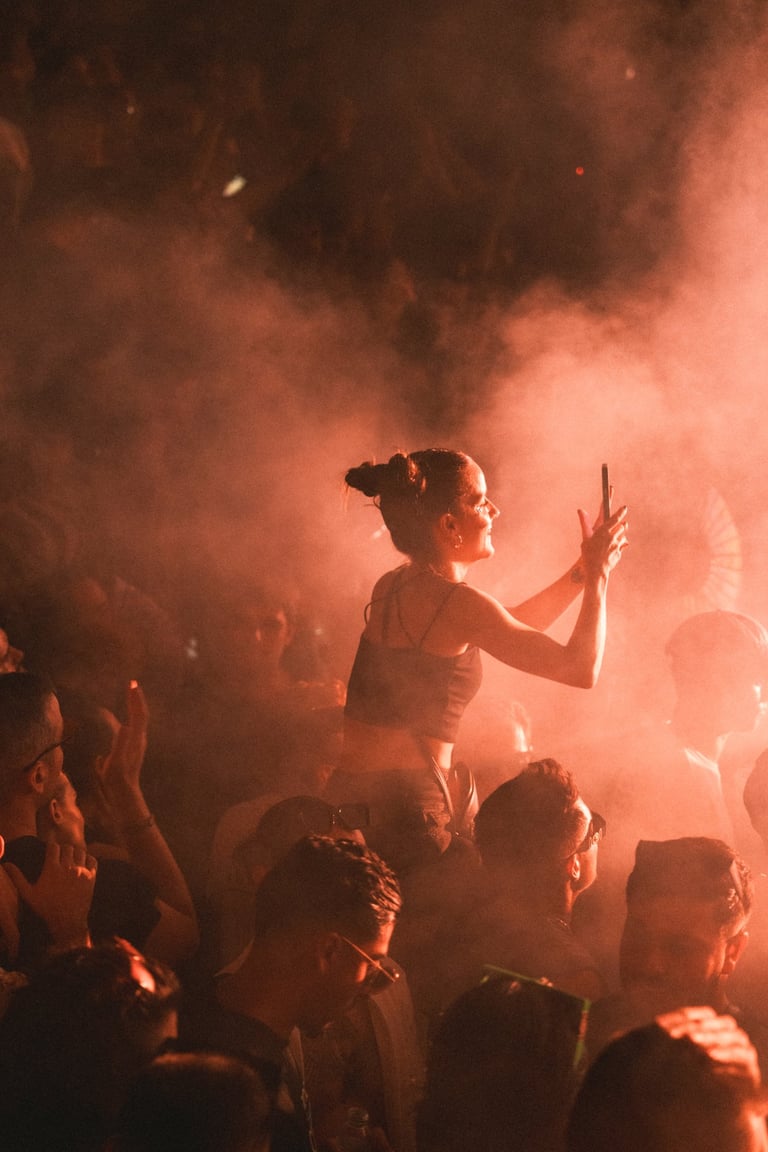 Woman on shoulders at a concert, filming with her phone. Smoky atmosphere with orange lighting. Energetic crowd. Live music e