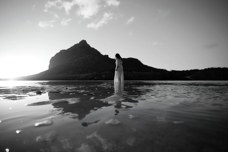 Woman standing in shallow water at sunrise near Le Morne Mountain in Mauritius, captured in dramatic black and white photo