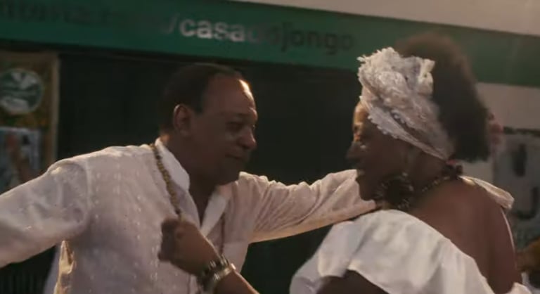 Homem e mulher negros dançam jongo na Casa Jongo, com roupas brancas e colares.