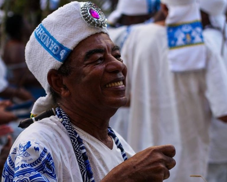 Foto de homem negro, sorridente, com traje branco e azul do bloco afro Filhos de Ghandi.
