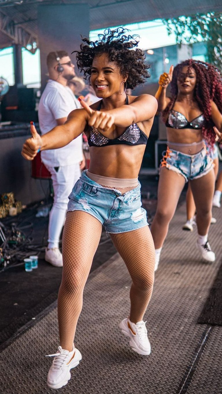 Duas mulheres negras dançam em ambiente festivo ao ar livre, vestindo top preto e short jeans.