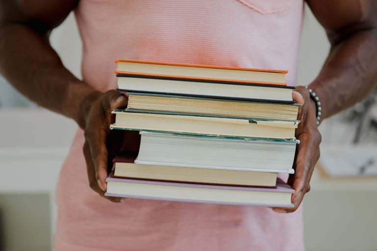 Foto de uma pessoa de pele retinta com camiseta rosa-claro segurando uma pilha de livros.