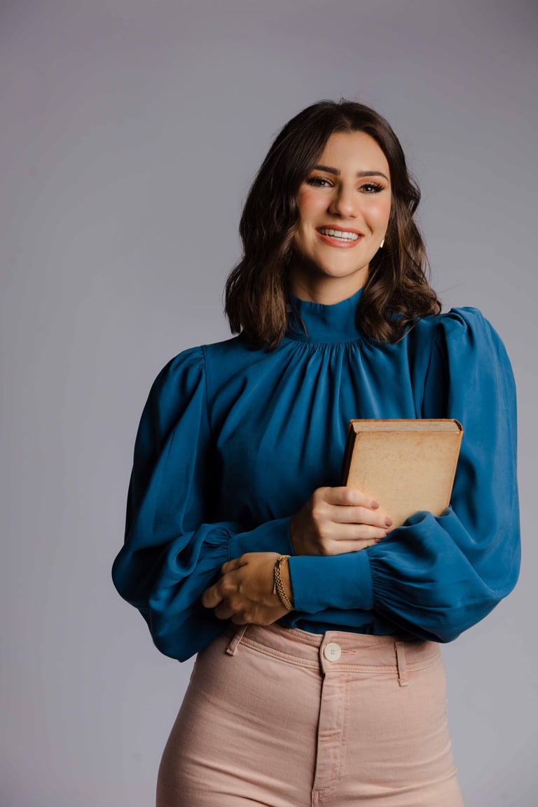 Smiling woman holding a vintage book wearing a blue silk blouse and pink trousers.