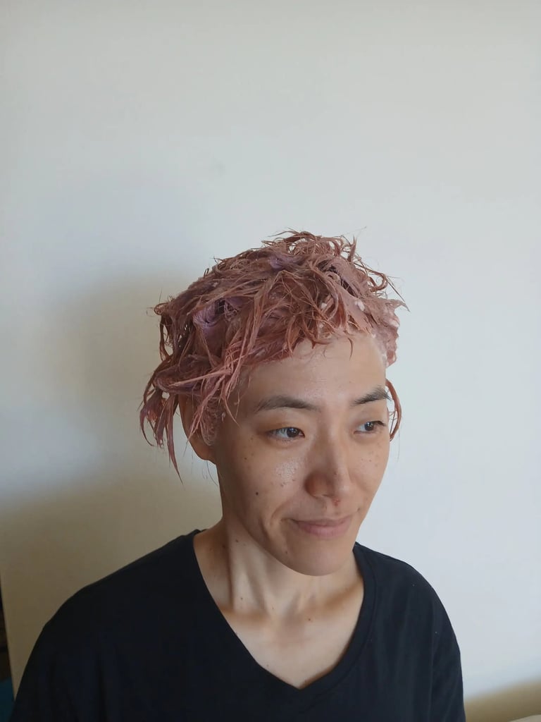 A person with short hair applying rose gold pink hair dye at home against a white wall.