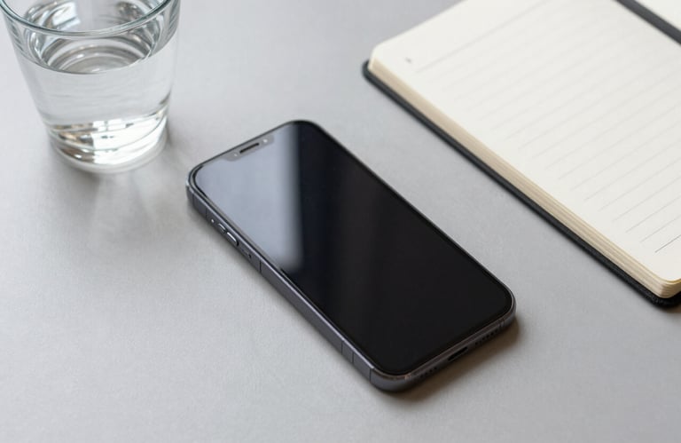 Photography from a high angle of a sleek mobile phone next to a glass of water and a notepad on a light gray desk, natural daylight, approachable and modern.