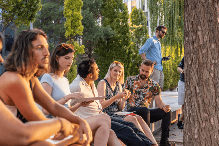 A croup of coworkers sitting on stairs at an event talking. Green trees in the background, sunlight hitting their faces.