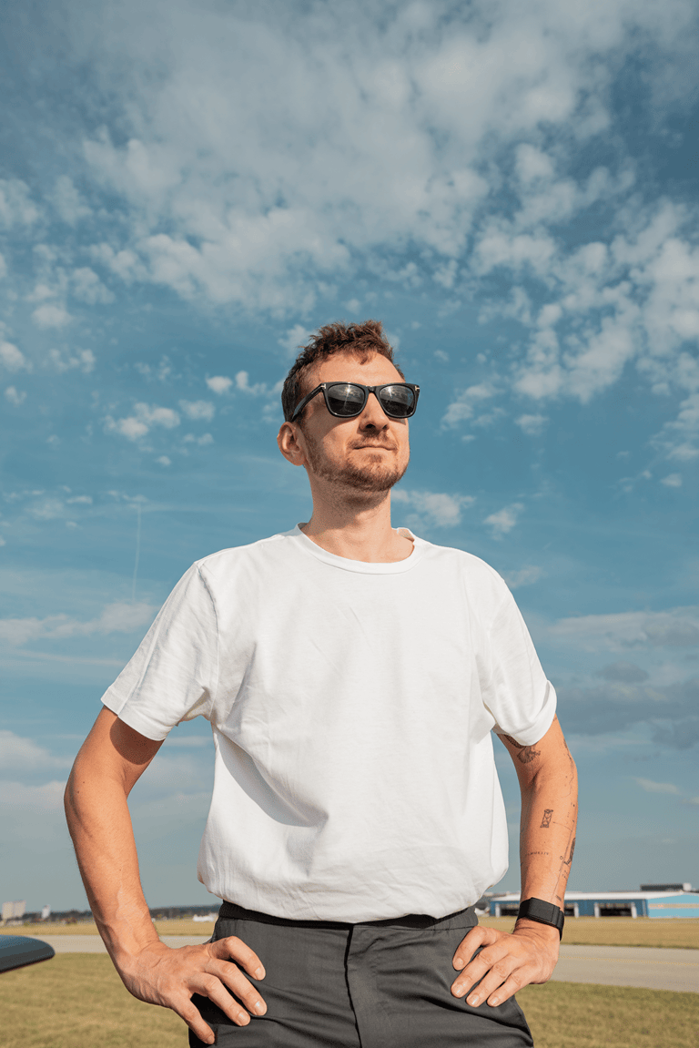 Young handsome man standing in the sun with sunglasses on. Hands placed on his hips, blue sky behind him.