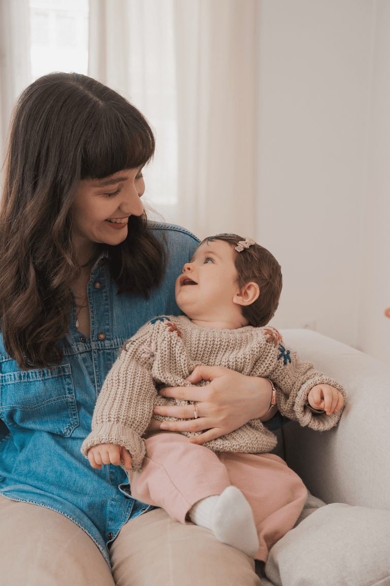 Young mom and baby sitting on a sofa and looking at each other, smiling.