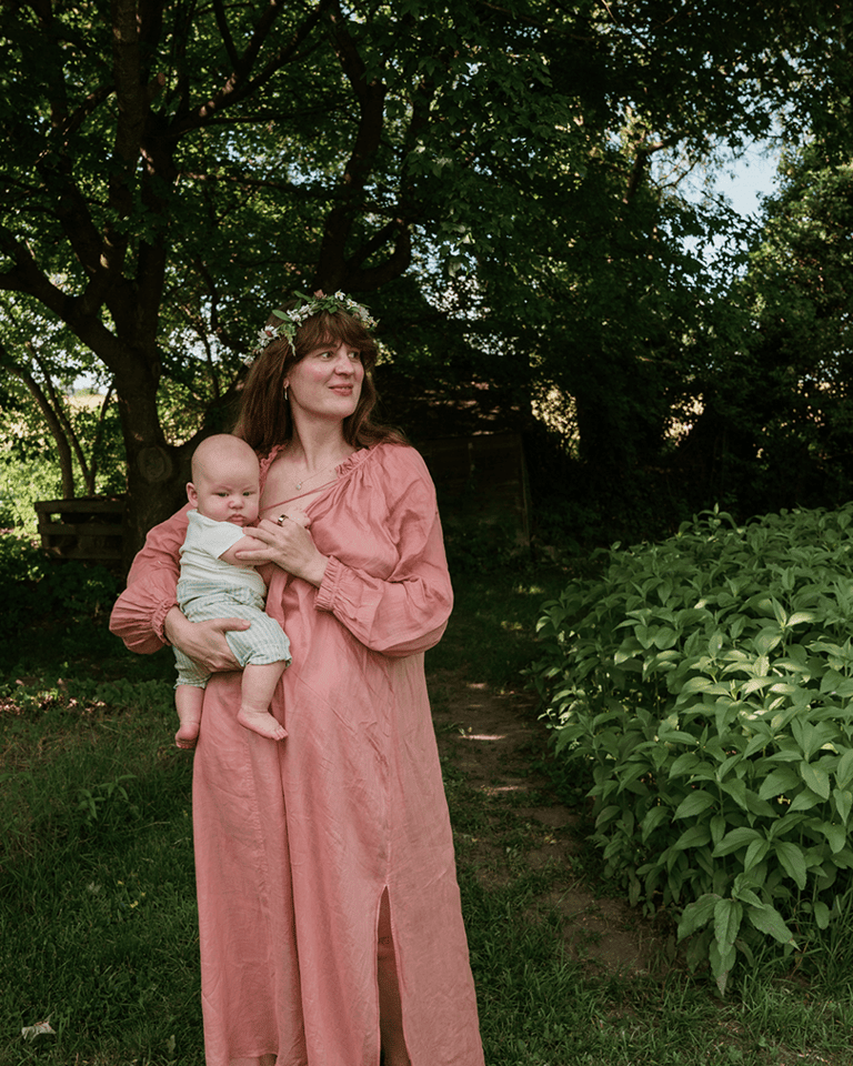 A woman standing surrounded by trees and green grass, smiling, looking away and holding her baby.
