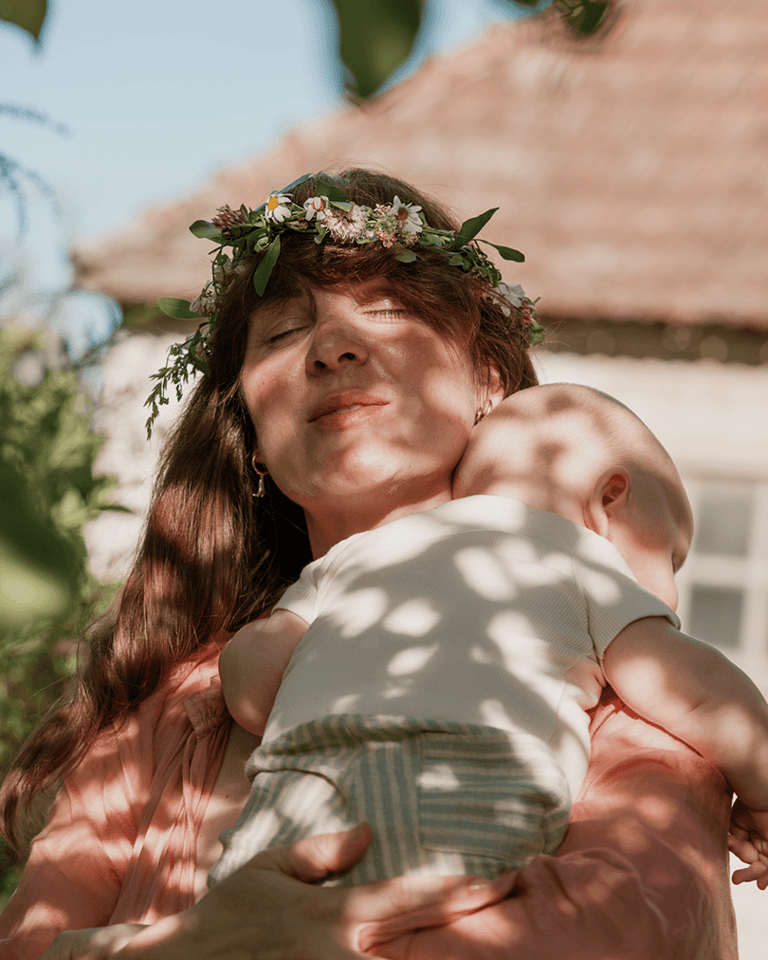 A mom holding her baby outside in the shade, sun shining through leaves that creates leafy shadows.