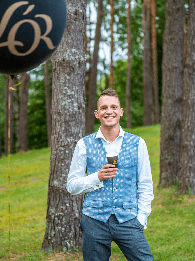 Young man standing in the forest with a cup of coffee looking into the camera and smiling.