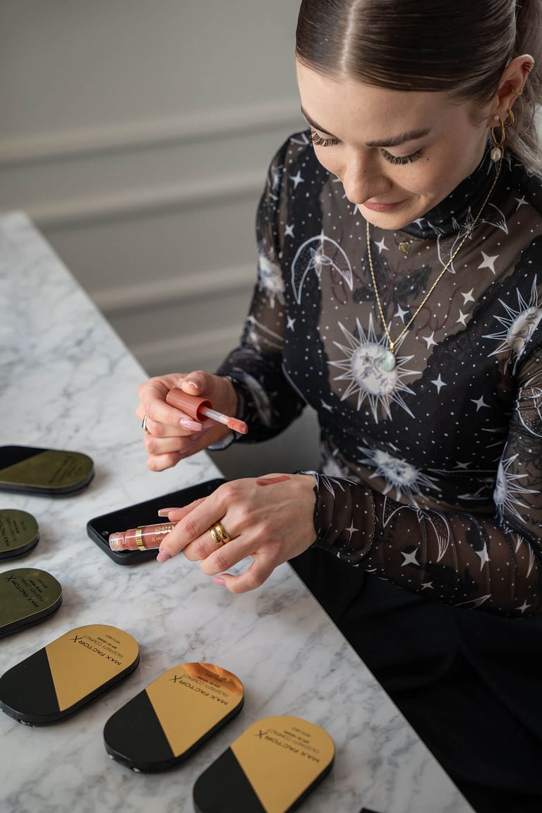 Young woman testing makeup on her hand, multiple pieces of makeup on the table in front.