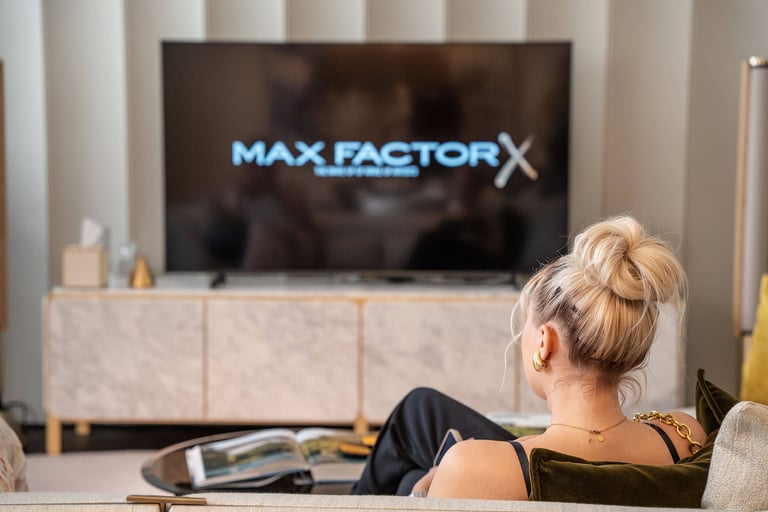 Influencer sitting on a sofa in front of a TV that has MaxFactor logo on it.