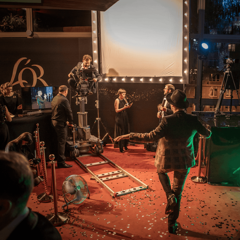 Red carpet event, man posing in front of a professional camera setup, bright lights and confetti.
