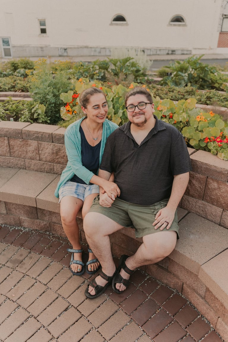 A smiling couple holding hands while sitting on a stone garden wall surrounded by lush flowers.