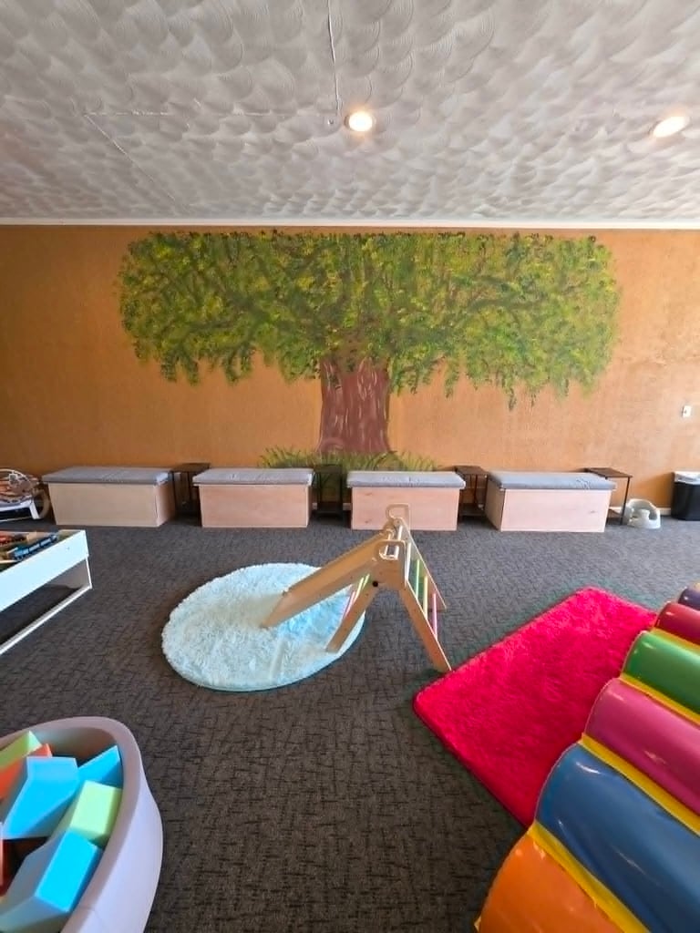 Indoor children's play area featuring a large tree mural, wooden benches, and colorful sensory toys.