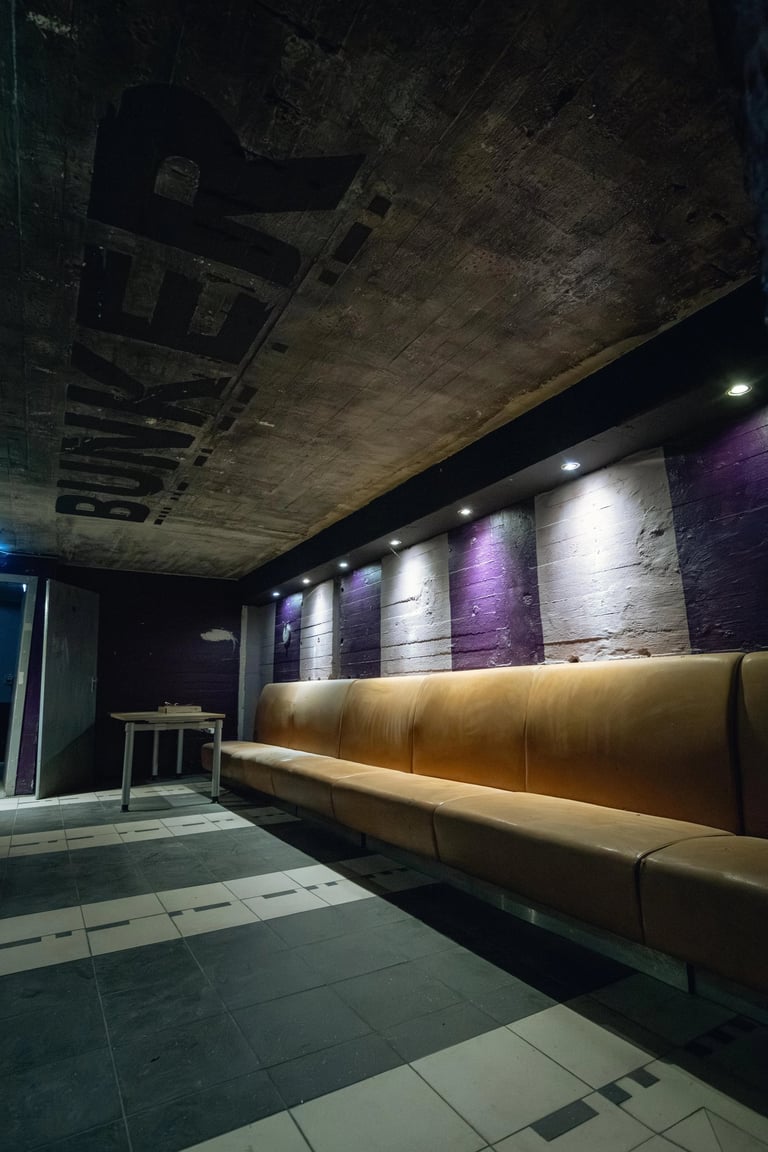 Underground bunker lounge featuring a long tan leather sofa against purple and white striped concrete walls.